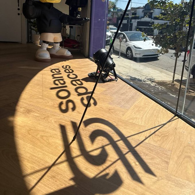Interior view through a glass window/door showing a wooden floor with 'Solar Space' text and logo, a stage light, a statue figure, and a white Tesla car parked on the street outside.