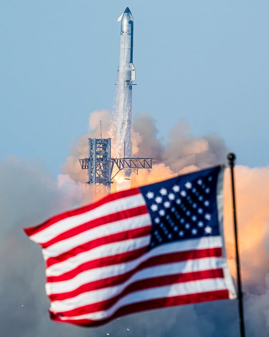 A SpaceX Starship rocket lifts off from its launch pad with bright exhaust plumes, while an American flag waves prominently in the foreground.