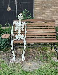 A life-sized plastic skeleton sitting upright on a wooden park bench in front of a brick building.
