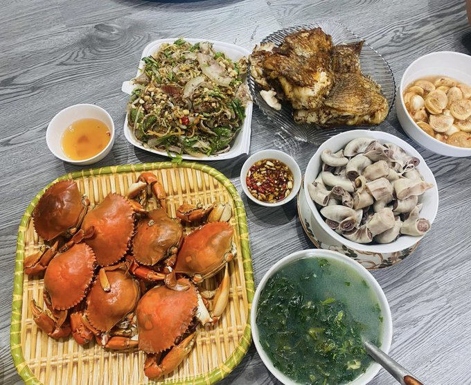 A table is filled with a feast including steamed crabs on a bamboo tray, grilled fish, a green salad, sliced boiled meat, dipping sauces, and a bowl of green soup.