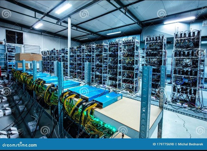 A large warehouse filled with rows of cryptocurrency mining equipment, featuring shelves of computer hardware and racks of wiring illuminated by overhead lights.