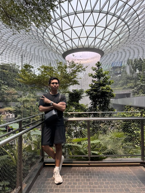 A man poses with crossed arms on a walkway at Jewel Changi Airport, with the iconic Rain Vortex waterfall cascading from the glass dome ceiling behind him, surrounded by lush indoor greenery. His caption "I'm inside you now Singapore! Where are you guys at?" playfully suggests he's being embraced by the stunning architectural marvel itself.