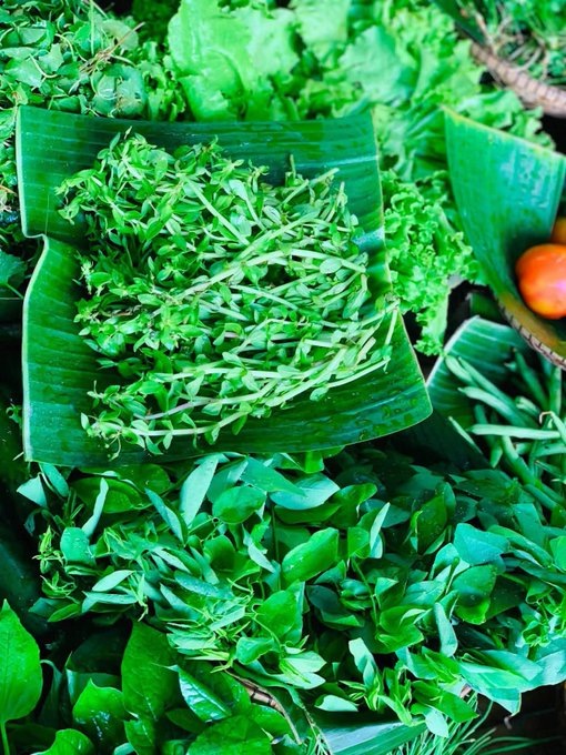Fresh green herbs and leafy vegetables displayed on banana leaves at a market, including what appears to be watercress and other greens.