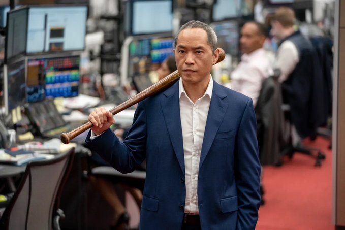 A serious-looking man in a navy blazer and white shirt carries a wooden baseball bat over his shoulder as he walks through what appears to be a busy trading floor, with computer monitors displaying financial data visible in the background.