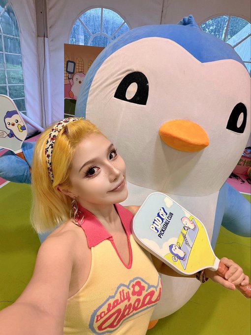 A young woman with blonde hair, a leopard print headband, and a yellow and pink top poses for a selfie next to a large blue and white penguin mascot, holding a paddle that reads 'TWT PINGU CLUB'.