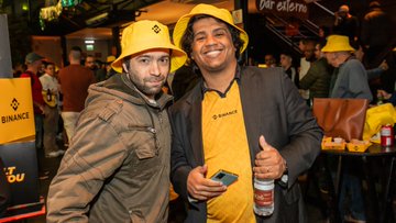 Two men in yellow Binance bucket hats pose together at a lively #BinanceTurns9 celebration, one wearing a branded Binance t-shirt and holding a drink, with the Binance logo visible on signage and merchandise in the background—capturing the festive spirit of the LATAM community gathering to mark the exchange's 9th anniversary.