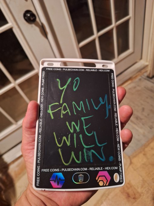 A hand holding a phone case designed like a chalkboard with handwritten green and blue text reading 'yo FAMILY WE WILL WIN,' bordered by text promoting PULSECHAIN.COM and HEX.COM with 'FREE COINS' and 'PUMP TIRES' slogans.