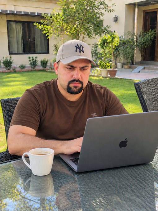 A man wearing a New York Yankees cap works on a gray Apple laptop at an outdoor table with a white coffee mug.