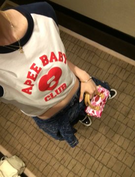 A person wearing a white "APEE BABY CLUB 8" ringer t-shirt and jeans, standing on a tiled floor while holding a small pink plush toy.