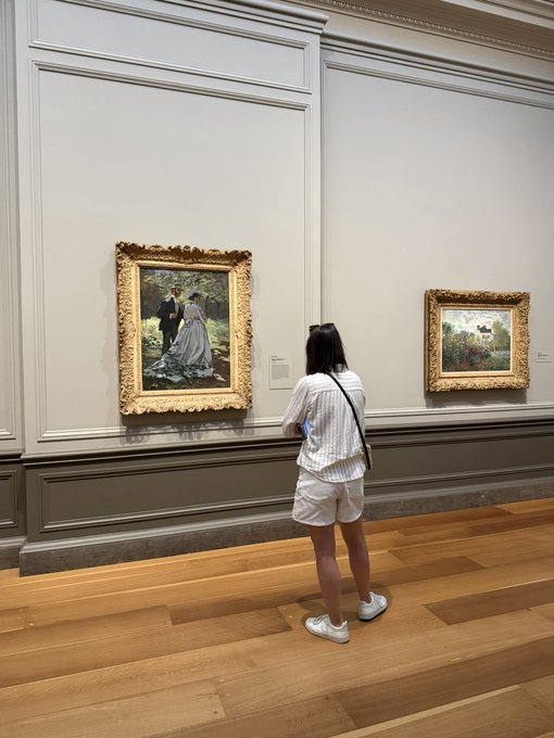 A person stands in an art museum gallery, viewing a framed Impressionist painting depicting a couple in a garden, with another landscape painting visible on the right wall.
