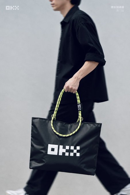 A person dressed in black walks while carrying a black Oxfam (OKX) tote bag featuring distinctive yellow and black braided rope handles.