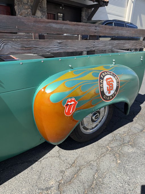 The rear fender of a vintage green vehicle decorated with orange and yellow flame decals, featuring a San Francisco Giants logo and a Rolling Stones tongue sticker.