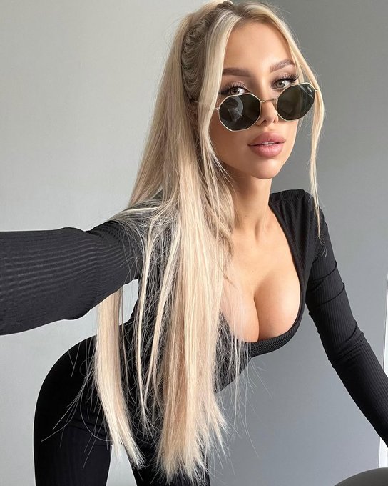 A selfie of a blonde woman wearing black round sunglasses and a black ribbed long-sleeve top with a deep neckline.