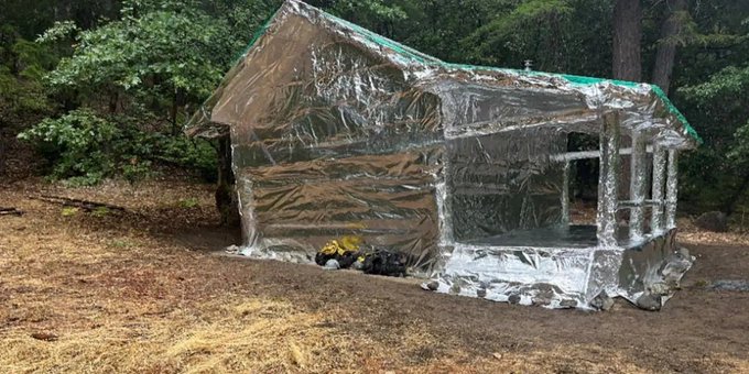 A backyard gazebo is wrapped entirely in silver emergency thermal blankets and secured with green tape, sitting on dry grass near some trees with camping gear on the ground in front.