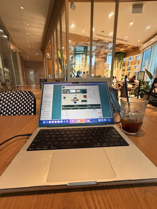 A laptop sits on a wooden table in a cafe or co-working space with an iced coffee beside it, showing what appears to be design or planning software on the screen.