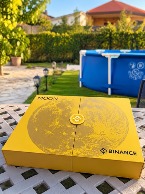 A yellow Binance-branded box labeled 'MOON' with a moon design, sitting on a white outdoor table with a blue Bestway pool in the background.