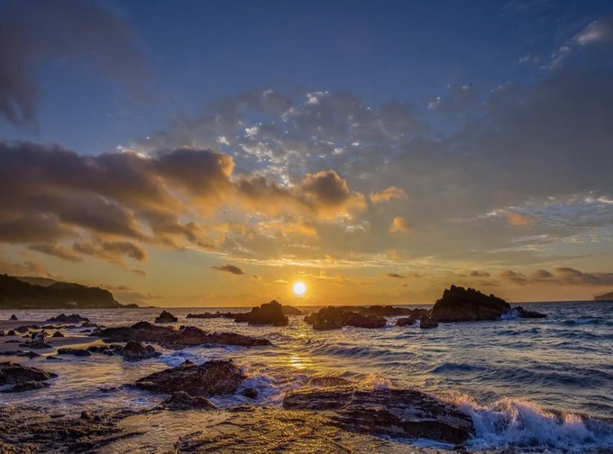 A scenic ocean sunset with the sun low on the horizon, casting golden light over rocky shoreline and gentle waves under a partly cloudy blue sky.