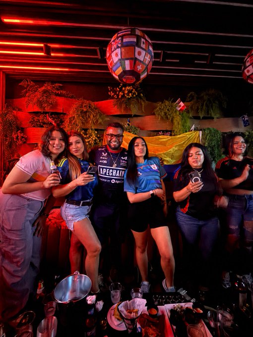 A group of friends pose together at a vibrant FIFA watch party, with several wearing Colombia jerseys and one man sporting an "APECHAIN" shirt, embodying the post's message of celebrating Colombian pride and ApeChain simultaneously under red-lit décor with international flag decorations.