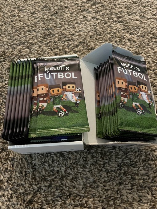 Two boxes of MEEBITS FUTBOL trading card packs displayed on a carpet, showing pixelated character soccer players on the green-themed packaging.