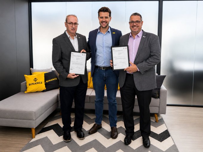 Three men in business attire pose holding framed ISO certification documents in a modern lounge area featuring "BINANCE" branded yellow pillows on a gray sectional sofa, celebrating Binance Brazil's newly earned ISO/IEC 27001 information security certification.