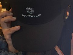 A close-up of a hand holding a black baseball cap with a white logo and the text 'MANTLE' on the front.