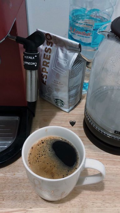 A white coffee cup containing black coffee with crema sits on a wooden counter next to an espresso machine with a milk frother and a bag of 'Espresso' coffee, with a water bottle visible in the background.
