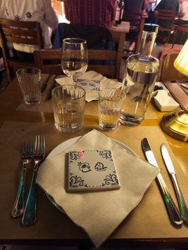 The image shows a table set for dinner with various glassware including wine glasses, cups, and water bottles. The table setting includes plates, silverware such as forks and knives, and a napkin featuring a picture of two faces on it. A bottle is also present on the table. The background reveals that this scene takes place in a restaurant or dining area. The post about dinner plans with friends in Lisbon seems to be related to the image, possibly suggesting an upcoming gathering or celebration involving people from Lisbon.