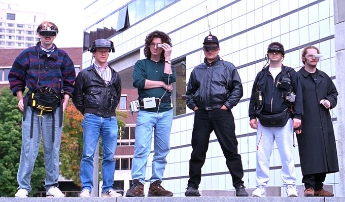 A group of six men wearing tactical headsets, visors, and various 1990s-style jackets and coats stand together in an urban setting with modern glass buildings in the background.