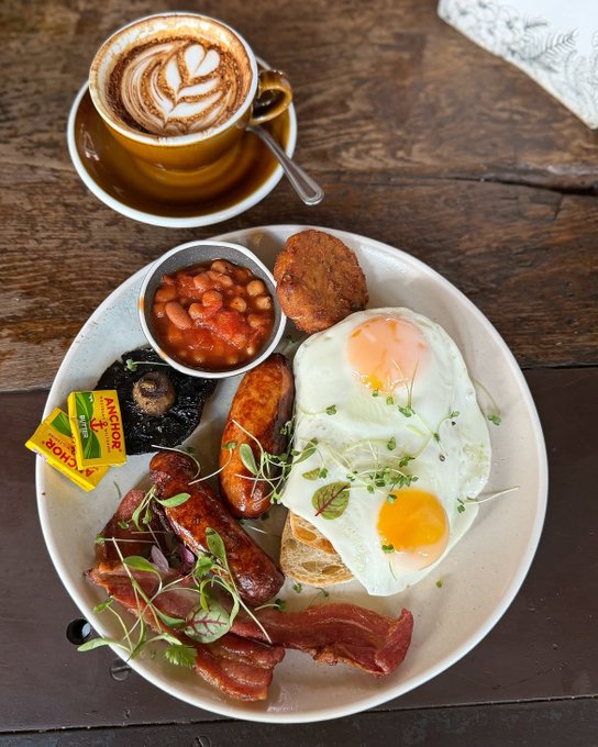 A traditional English breakfast plate featuring fried eggs, bacon, sausage, baked beans in a small ramekin, a hash brown, a grilled portobello mushroom, and microgreens, served alongside a latte with latte art in a brown cup on a wooden table.