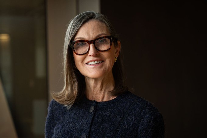 A woman wearing glasses and a navy blue sweater stands in front of a window and poses for the camera. The accompanying text says "Cathie Wood urges investors to sell gold for Bitcoin", which suggests that the woman's opinion about Bitcoin has influenced her advice on investing. She advises selling gold and buying Bitcoin, expecting it to reach $1,500,000 within a year, based on her knowledge or experience in the matter.