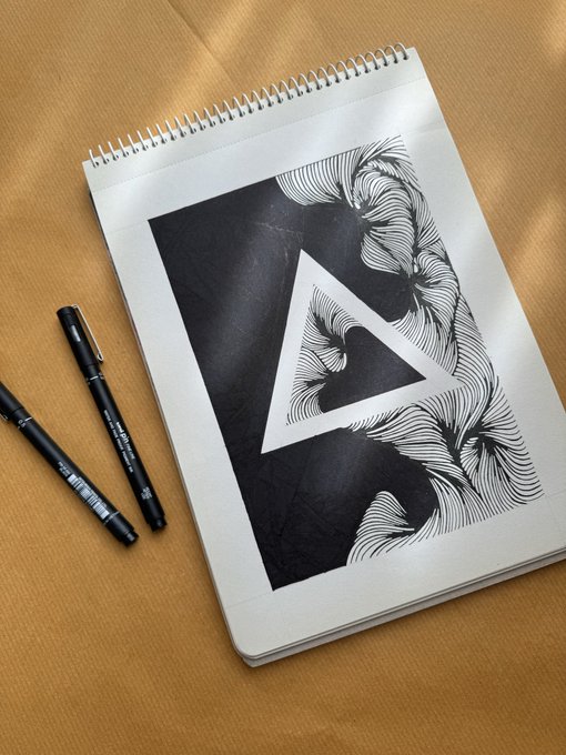 The image features a sketchbook with a black and white drawing of a triangle, accompanied by two pens - one black and one black and blue. The sketchbook has a spiral binding, which adds an artistic touch to its appearance. The pen on the left is black, while the pen on the right is black and blue. These pens are placed next to each other, with their tips pointing towards the triangle drawing in the sketchbook.