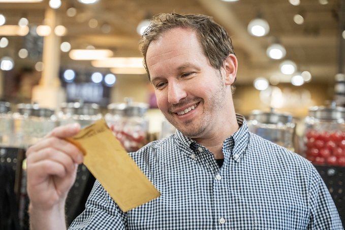 A smiling man in a blue checkered shirt holds up a small brown paper bag in what appears to be a candy or bulk goods store with jars visible in the background.
