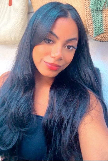 A selfie showing a smiling woman with long dark hair and red lipstick, wearing a black top, with woven baskets visible in the background. The image aligns with the post's "happy Saturday friends ❤️" greeting, conveying a cheerful, ready-to-start-the-day vibe.