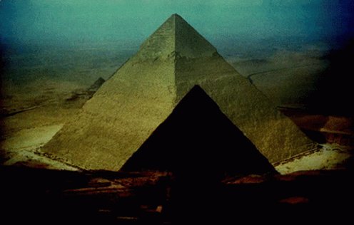 The image features a pyramid-like structure, possibly an ancient Egyptian pyramid or a pyramid-shaped building, situated in a desert setting with a blue sky above. The pyramid appears to be made of stone, giving it a historical and majestic appearance. The surrounding area seems barren, with no visible vegetation or other structures nearby.
