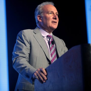 Peter Schiff stands at a podium, mid-speech, delivering remarks on Bitcoin and his personal stance on forgoing the cryptocurrency over the past five years.

Image: A man with graying hair speaks at a podium, his hand resting on the edge, against a dark blue-lit background.