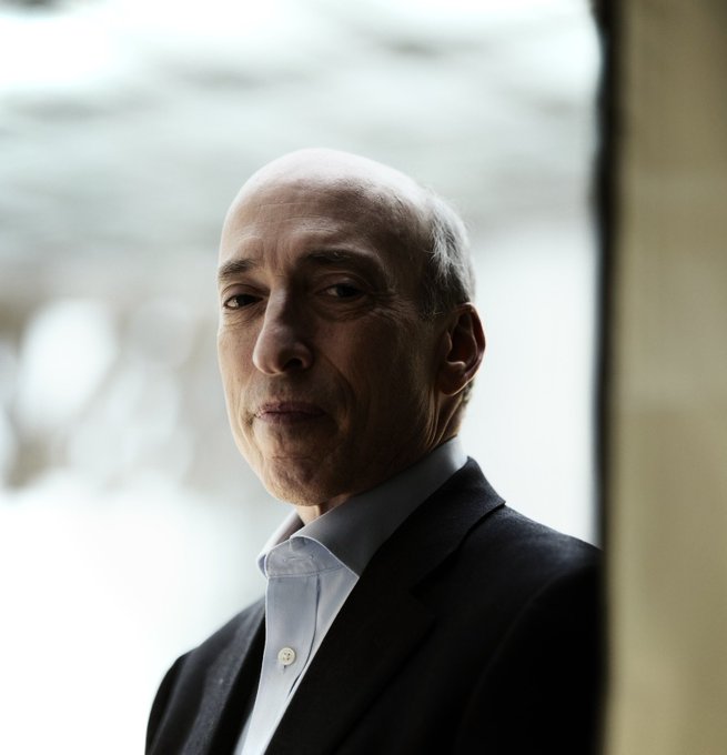 A portrait of a bald older man in a dark suit jacket and light blue collared shirt, standing against a light-colored background.