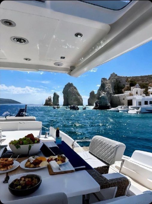 A view from a boat dining area featuring a spread of food on a table in the foreground, with the famous Faraglioni rock formations of Capri rising from the turquoise sea in the distance under a sunny sky.