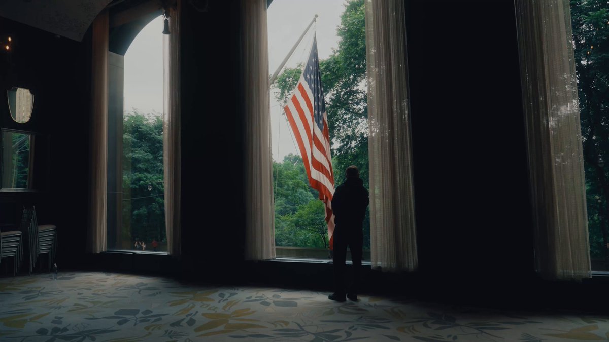 The image shows a man standing near a window, holding an American flag that has three vertical stripes of red, white, and blue colors. The scene appears to be indoors with a dining table nearby. The man seems to be looking out the window or observing something outside while still keeping his focus on the flag. The post about the building sushi in public is related to this image as it suggests an interesting juxtaposition between the traditional flag of the United States and the modern concept of sushi, which could symbolize a blend of cultural elements or a commentary on the changing times.