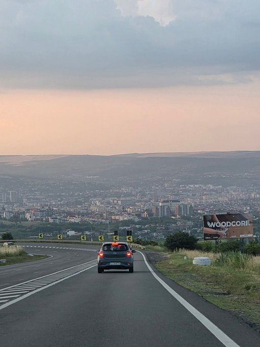 A car drives along a highway overlooking a sprawling city at dusk, with a "WOODCORE" billboard visible on the right side of the road.