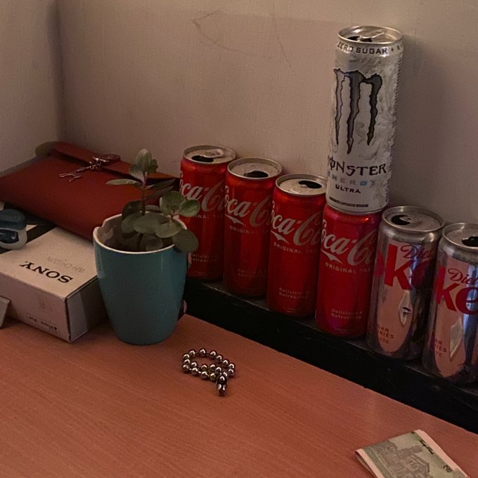 A photo of a desk with a small potted plant, a Sony box, several empty Coca-Cola and Diet Coke cans, a Monster Energy Ultra can, a red purse, and some paper currency.