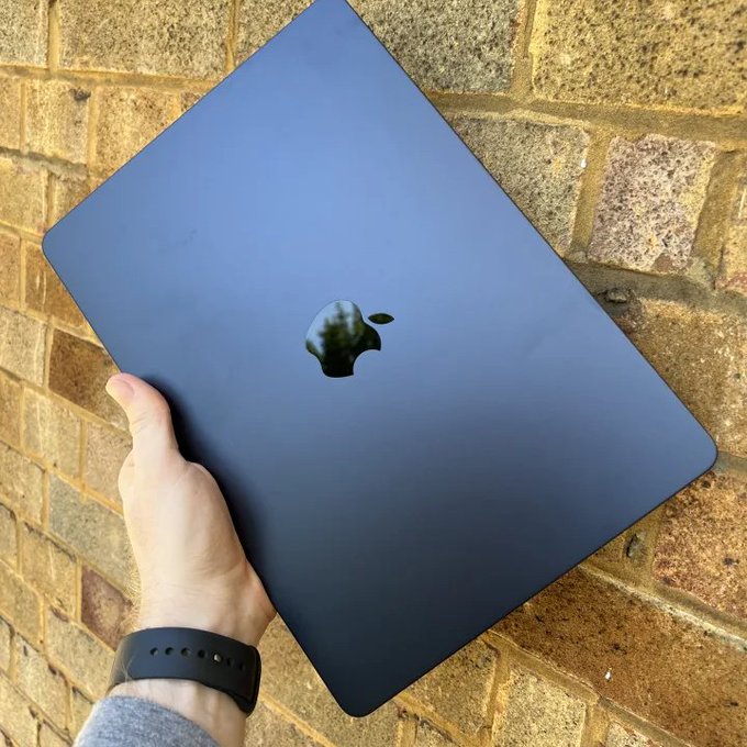 A hand holding a black/dark blue Apple MacBook closed, showing the Apple logo on the lid, against a stone brick wall background.