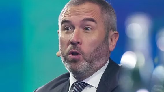 Brad Garlinghouse, Ripple's CEO, is shown speaking with an animated expression while wearing a dark suit and tie with a small headset microphone, set against a blurred blue and teal background. The image accompanies news about Ripple's projected $1 billion revenue run-rate target for 2026, a key milestone under his leadership.