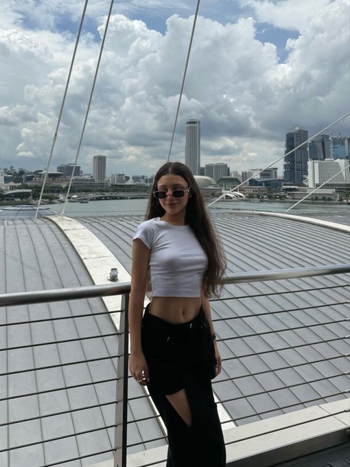 A young woman in a white crop top, black pants, and sunglasses poses against a city skyline, captioning her post to announce she's packing for TOKEN2049 and inviting content collaborations from projects and fellow creators.