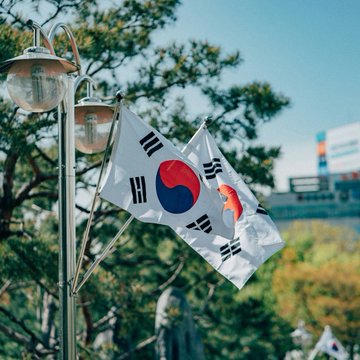 Two South Korean flags flutter on a lamppost against a backdrop of green trees, symbolizing the nation as it prepares to roll out its three-phase roadmap to tokenize stocks, bonds, and funds beginning in early 2027.