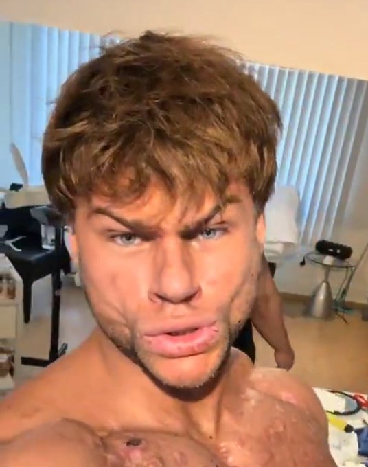 A close-up selfie of a young man with tousled brown hair and light facial hair, shirtless, in a room with vertical blinds and furniture in the background.