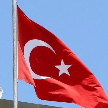 Photograph of the Turkish flag (red with a white crescent and star) waving on a flagpole against a clear blue sky.