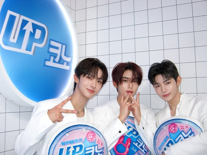 Three young men in white outfits pose with peace signs in front of a large blue and white "UP" logo, holding round promotional discs branded with "업코노" (UpKono) graphics. The image accompanies a behind-the-scenes post for EP.14 of UP CONO, teasing AND2BLE's release on July 23, 2026 at 7PM KST.