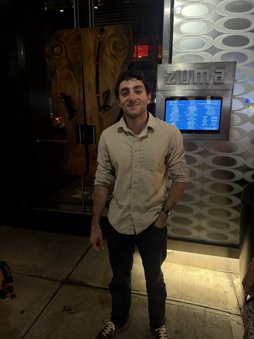 A man in a beige button-down shirt and jeans stands smiling outside "Zuma," a restaurant whose illuminated logo and digital menu board are visible behind him, suggesting the social post is speculating whether he could be the winner of the Solana poker tournament spotted at this upscale venue.
