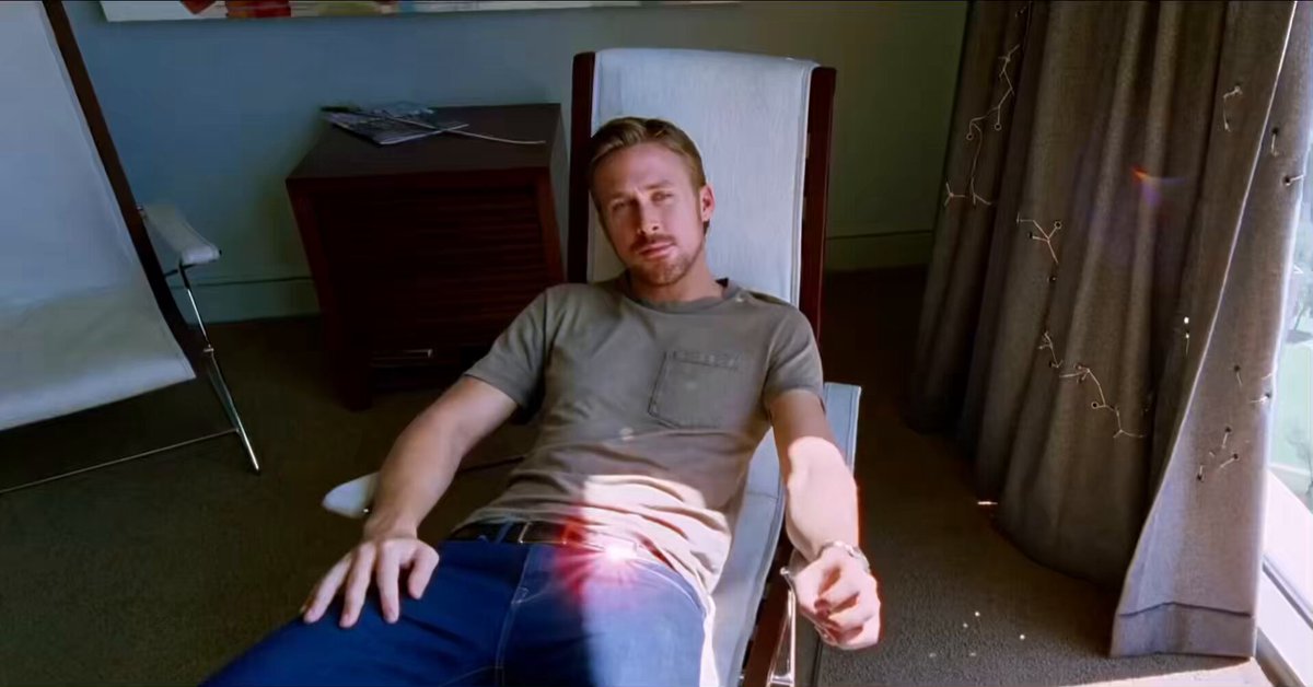 In this scene, a man is sitting comfortably in a chair in front of a window. The room has beige carpeting and white walls, creating a calm atmosphere. Behind him, there's a wooden bed frame with a striped headboard that adds to the room's overall style. A desk with a chair can also be seen nearby. The man appears to be looking at something outside the window or engaging in an activity related to the post displayed on the wall about comparing prices for Bitcoin.