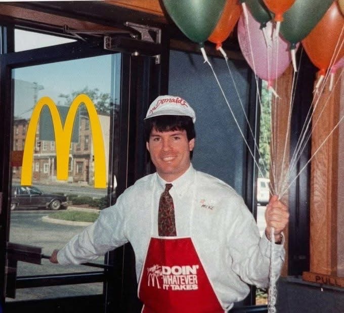 The image features a man wearing an apron with "Doin' What Ever Works" written across his chest, standing next to a McDonald's restaurant. He is holding balloons that are tied together, creating a festive atmosphere around him. The post in the image reads "Here comes the diamond hands", which could be interpreted as a reference to the man's job at the restaurant or simply an interesting phrase used for decoration purposes.

The scene takes place inside a McDonald's restaurant, with a dining table and chairs visible in the background. There are also two cars parked outside the restaurant, one on each side of the image.